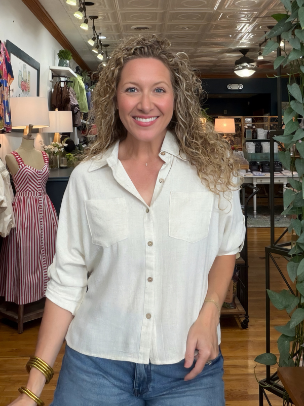 A smiling woman with curly blonde hair wearing a light cream linen button-down shirt and blue jeans, standing inside a boutique store.