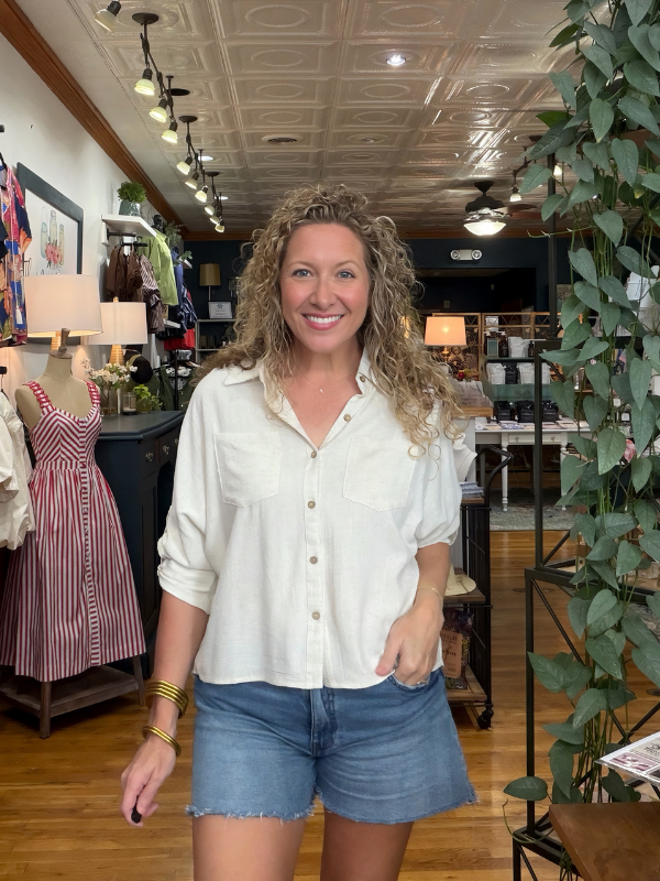 A smiling woman with curly blonde hair stands inside a boutique shop wearing a cream-colored, long-sleeve button-down shirt and denim shorts.