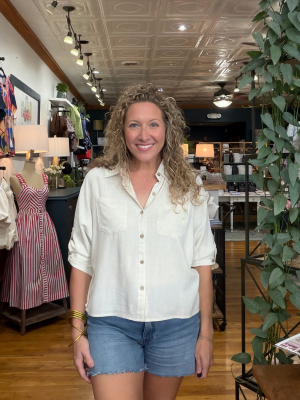A smiling woman with curly blonde hair stands inside a clothing boutique, wearing a cream-colored linen button-up shirt with rolled sleeves and blue denim shorts.
