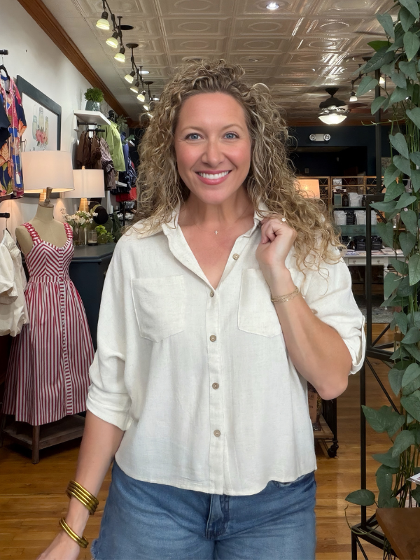 A smiling woman with curly blonde hair stands inside a clothing boutique wearing a light cream-colored linen button-up shirt and blue jeans.