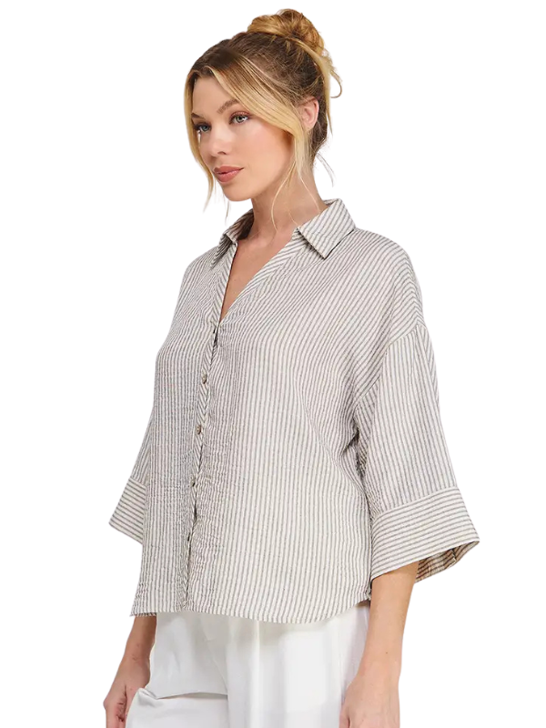 A woman wearing a loose, short-sleeve button-up shirt with vertical beige and white stripes, paired with white pants, photographed against a solid white background.