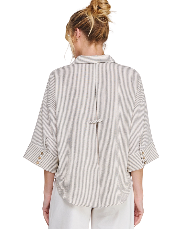A woman shown from the back wearing a loose-fit, off-white shirt with thin vertical gray stripes, featuring three-quarter sleeves and a tie detail at the center back. She is also wearing light-colored trousers, set against a plain white background.