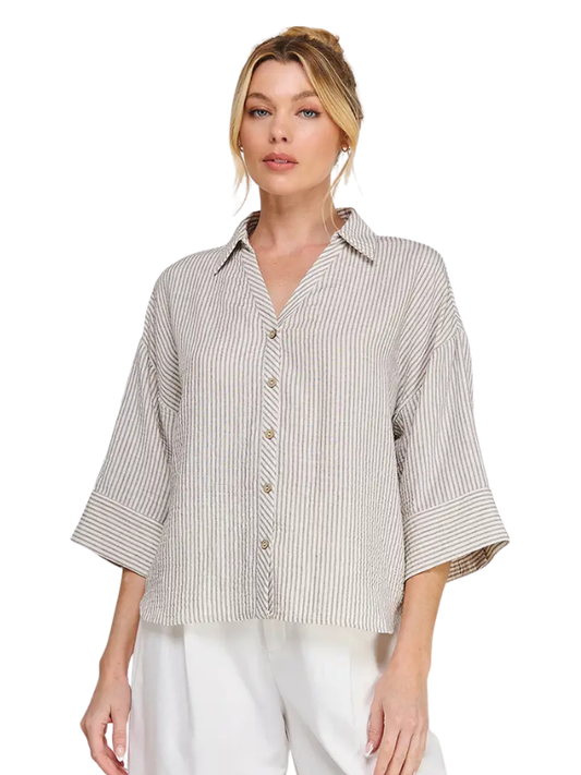 A woman wearing a beige and white vertically striped button-down shirt with three-quarter sleeves, paired with white pants, against a plain white background.