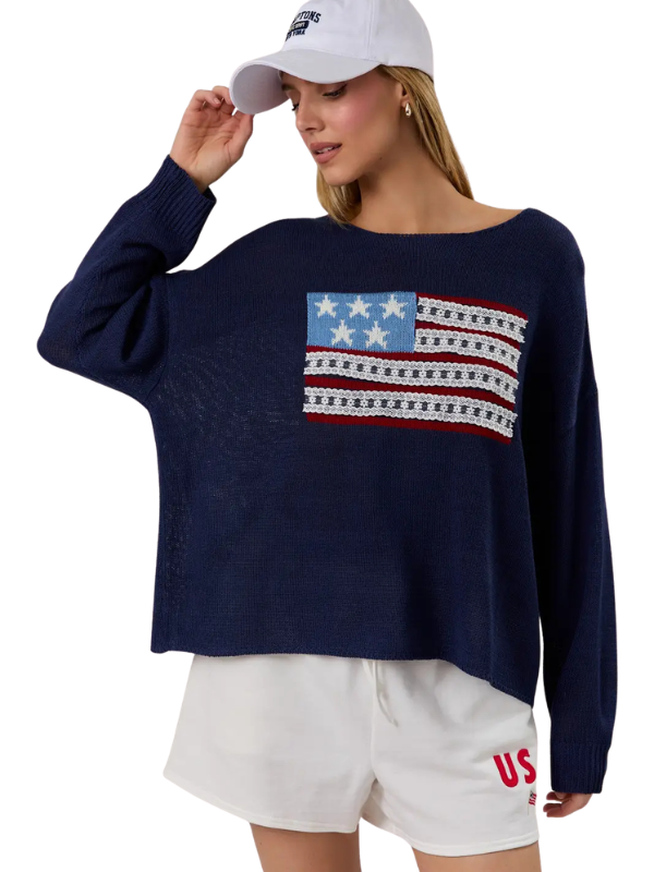 Woman wearing a navy sweater with an American flag design, white shorts, and a white cap on a white background.