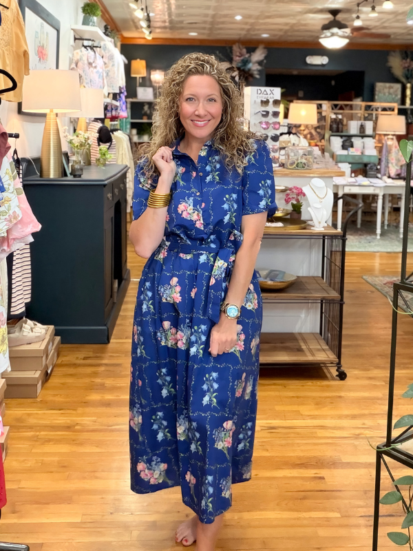 Woman in a blue floral dress standing in a store with wooden floors and various items on display.