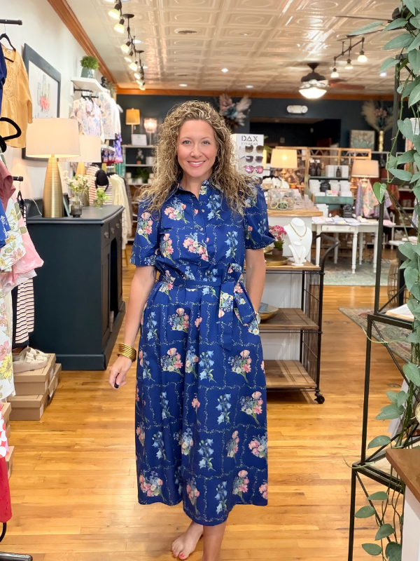 Woman in a blue floral dress standing in a store with wooden floors and shelves.