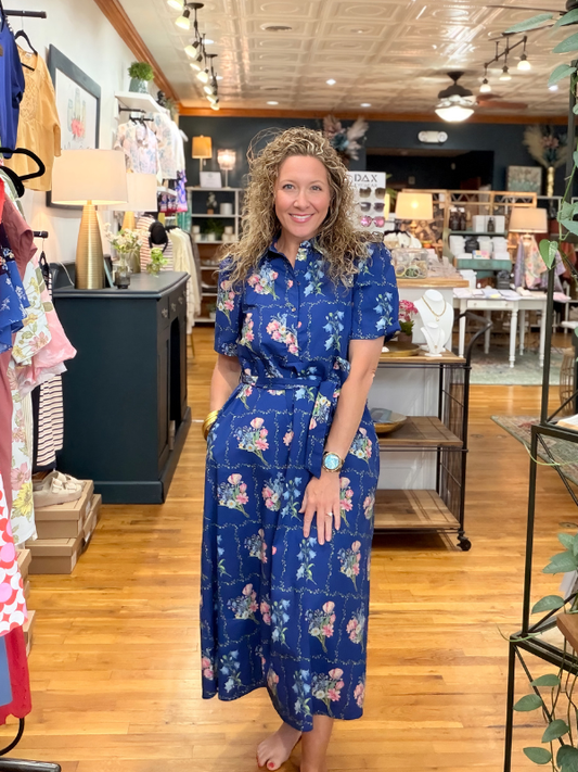 Woman in a blue floral dress standing in a store with wooden floors and shelves.