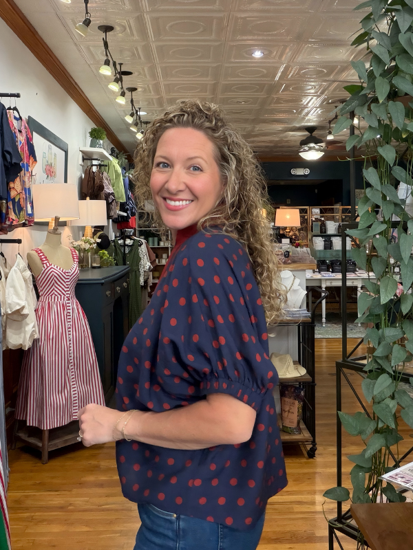 A smiling woman with curly hair stands in a boutique shop, wearing a navy blue blouse with red polka dots and blue jeans.