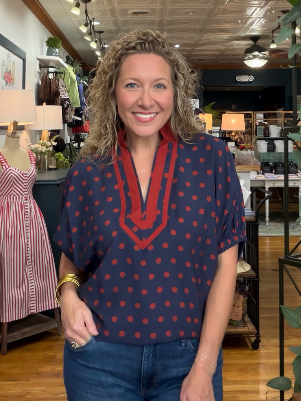 A smiling woman with curly blonde hair stands in a boutique wearing a navy blue short-sleeve top with red polka dots and a red V-neck trim, paired with blue jeans.
