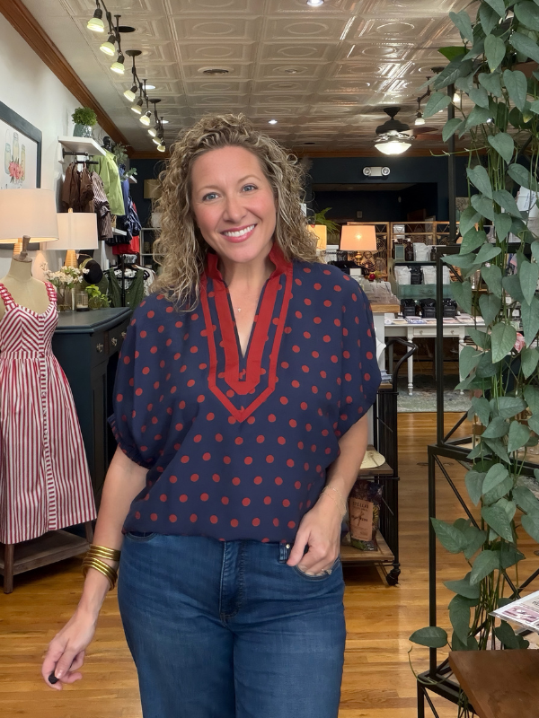 A woman with curly blonde hair stands in a boutique wearing a navy blue short-sleeve top with red polka dots and a red V-neck trim, paired with blue jeans.