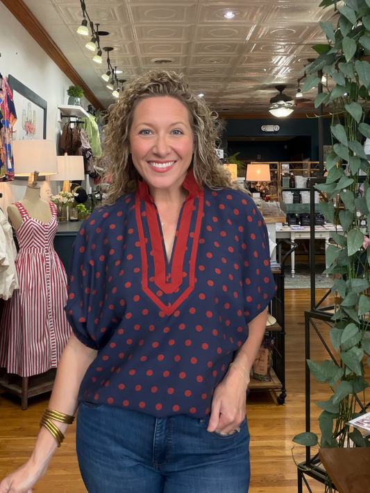 A smiling woman with curly hair stands inside a boutique wearing a navy blue short-sleeve blouse with red trim and red polka dots, paired with blue jeans.