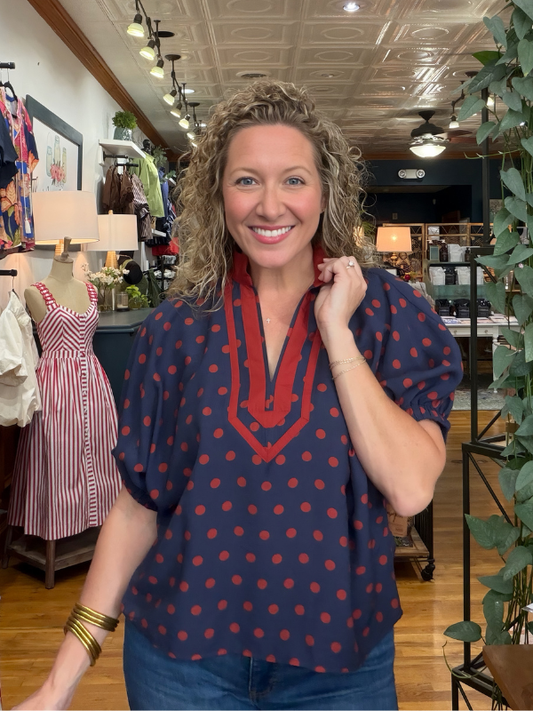 A smiling woman with curly blonde hair models a navy blue blouse with red polka dots and a red V-neckline detail, standing inside a boutique store.