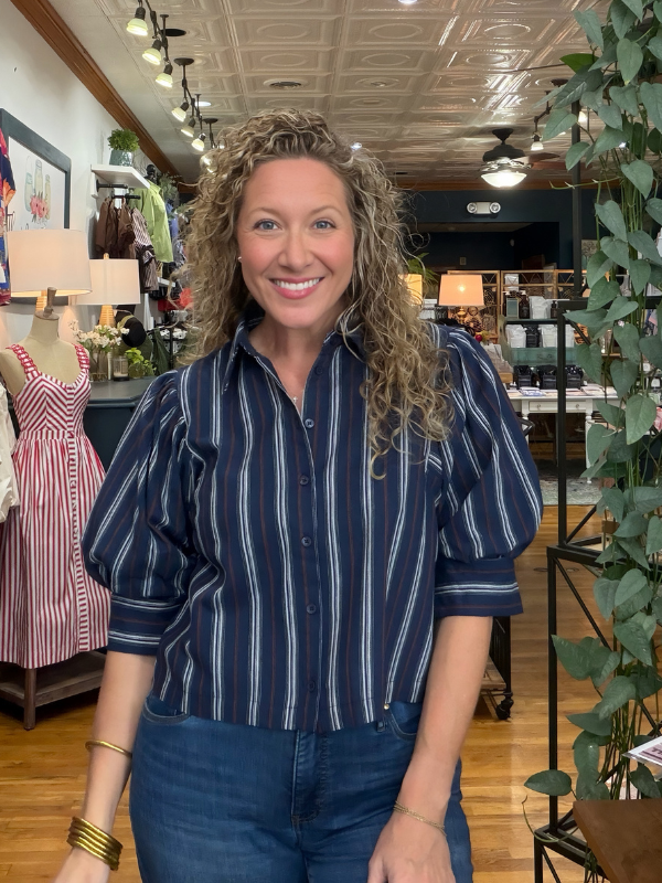 A smiling woman with curly hair stands in a boutique wearing a navy blue button-up shirt with vertical white and thin gold stripes, paired with blue jeans.