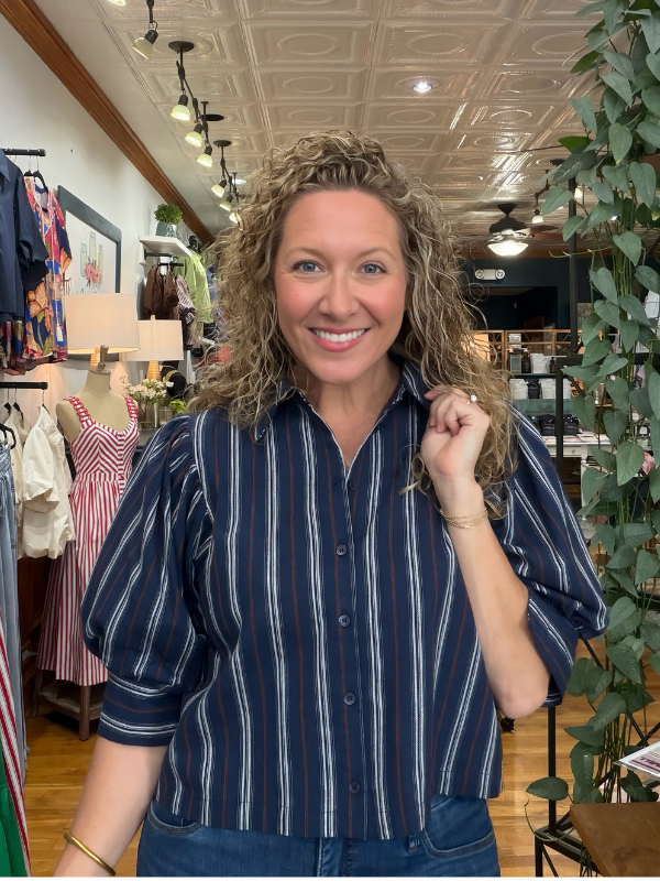 A smiling woman with curly hair wearing a navy blue button-down shirt with thin white vertical stripes, standing inside a boutique store.