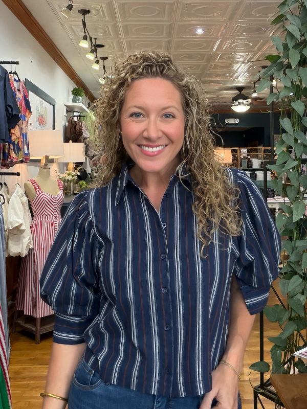 A smiling woman with curly blonde hair wearing a navy blue button-up shirt with vertical white and thin red stripes, standing inside a retail boutique.