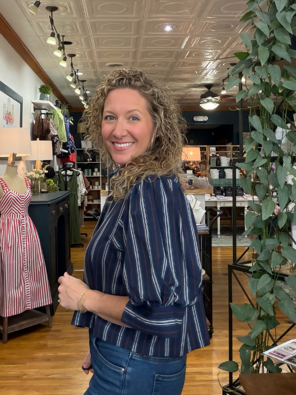 A smiling woman with curly hair stands in a boutique shop wearing a navy blue and white vertically striped blouse with puff sleeves and blue jeans.
