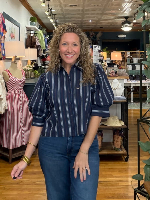 A smiling woman with curly hair stands in a clothing boutique wearing a navy blue and white vertically striped button-down shirt with puff sleeves and blue jeans.
