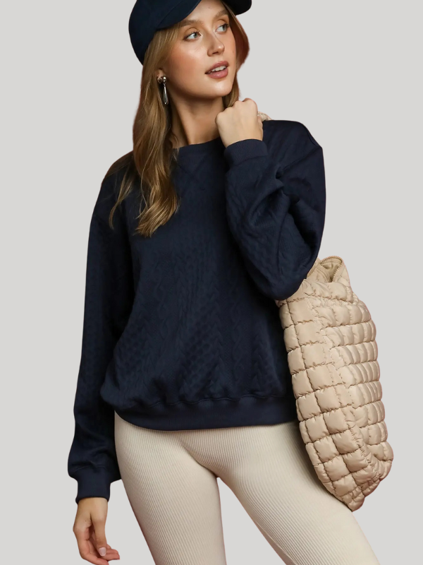 Woman wearing a navy sweater and cap, holding a beige quilted bag against a light background