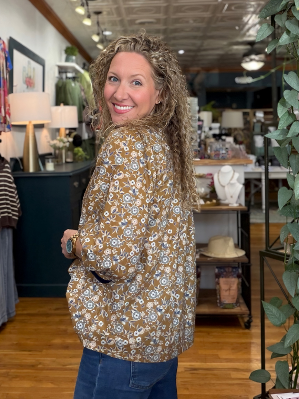 A woman with curly blonde hair wearing a brown long-sleeve jacket with a white and blue floral pattern, standing in a boutique store.