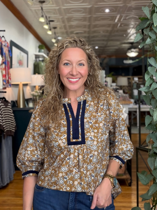 A smiling woman with curly blonde hair wearing a brown and white floral patterned blouse with navy blue trim, standing inside a boutique.