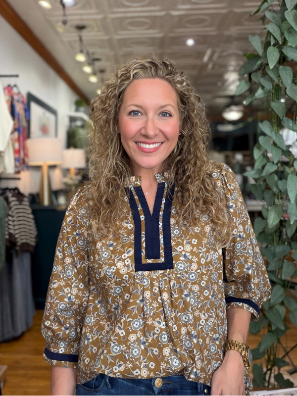A smiling woman with curly blonde hair wearing a brown and blue floral patterned blouse with a navy V-neck trim, standing inside a boutique.