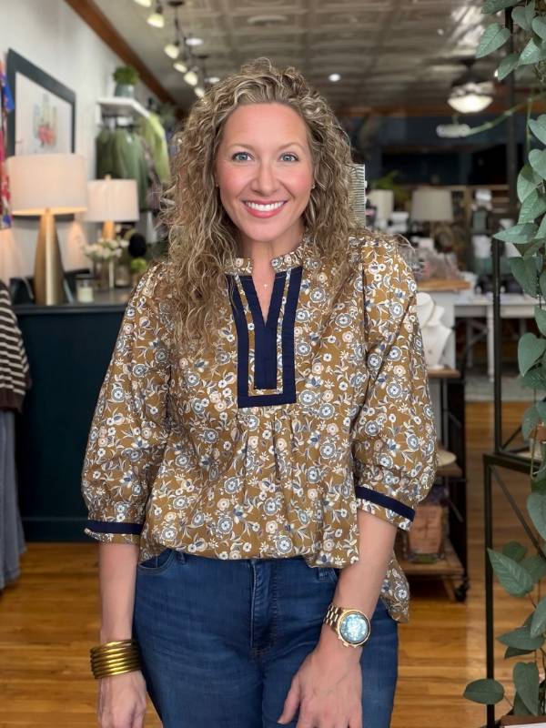 A smiling woman with curly blonde hair stands in a boutique wearing a brown and beige floral print blouse with navy blue trim, paired with blue jeans.