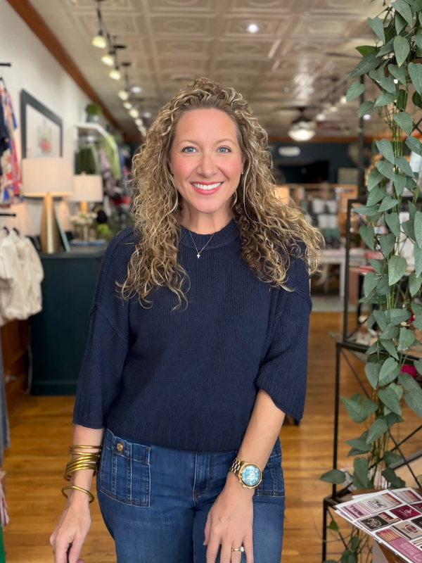 A smiling woman with curly blonde hair stands in a boutique wearing a dark navy blue ribbed knit sweater and blue jeans. She is accessorized with gold bracelets, a watch, and a necklace.