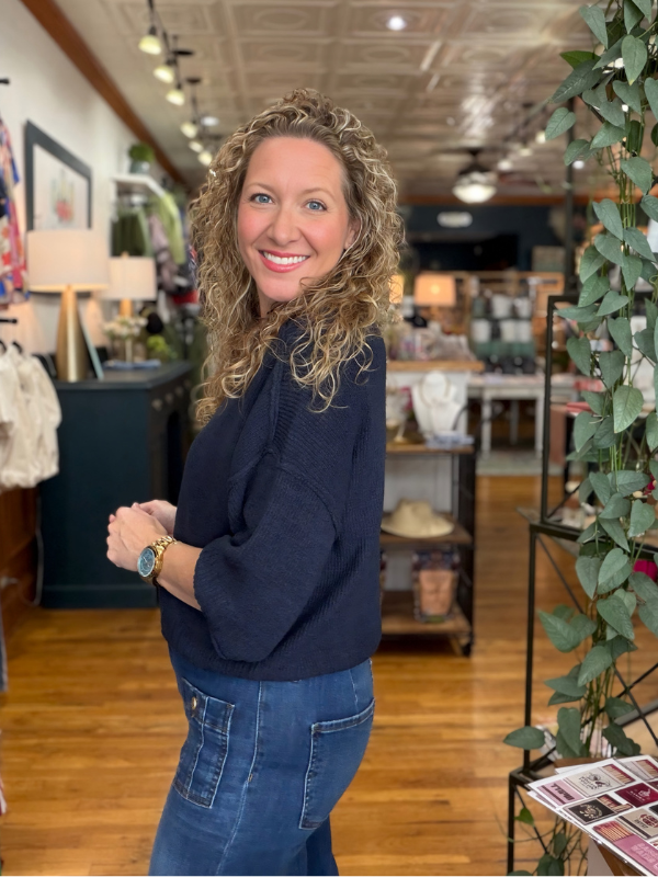 A smiling woman with curly blonde hair stands in a boutique shop, wearing a dark navy blue knit sweater and blue jeans.