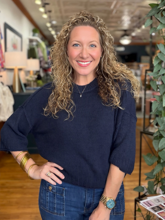 A smiling woman with curly blonde hair wearing a dark navy blue ribbed knit sweater and blue jeans, standing in a retail store interior.