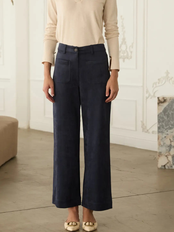 A model wearing navy blue wide-leg cropped trousers with a beige long-sleeve top and gold flat shoes, standing in a bright, elegant room with white paneled walls.