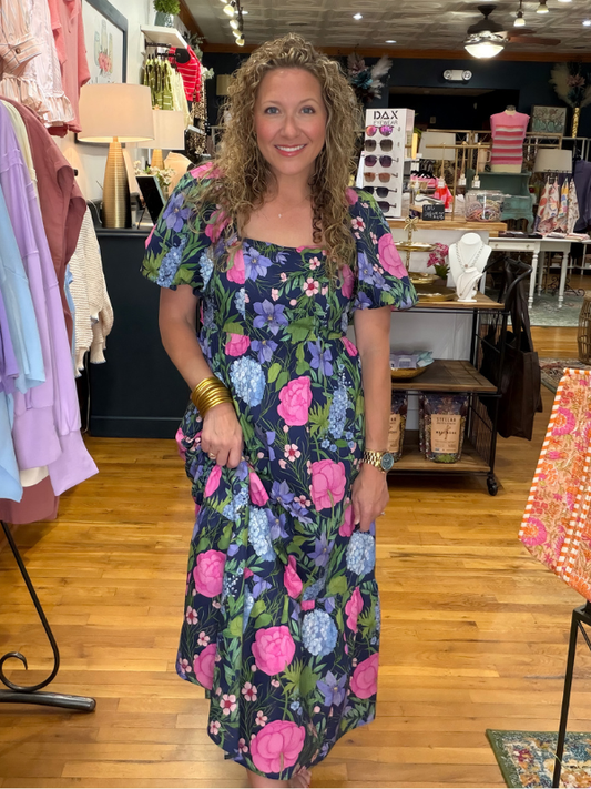 Woman in a floral dress standing in a store with clothing and accessories in the background.