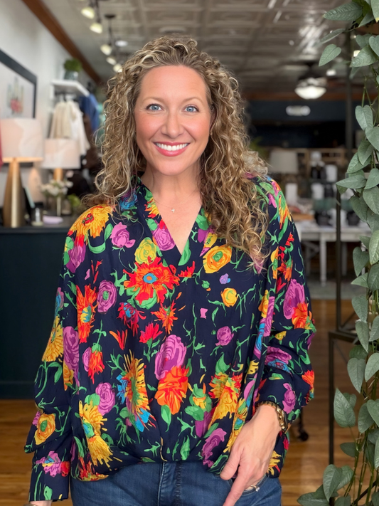 A smiling woman with curly blonde hair wearing a vibrant, multicolored floral print blouse with blue jeans, standing in a boutique setting.