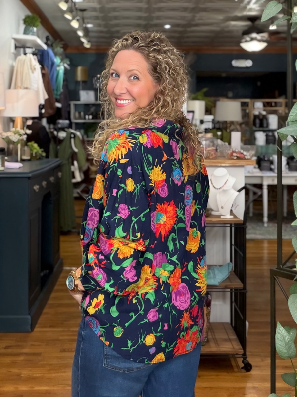 A woman with curly blonde hair stands in a boutique shop, wearing a navy blue long-sleeve blouse with a vibrant multicolor floral print and blue jeans.