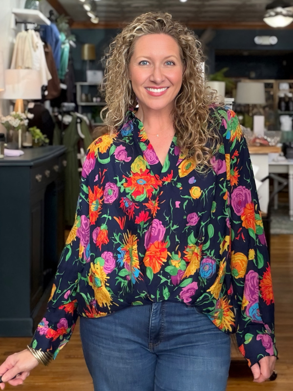 A smiling woman with curly blonde hair wearing a long-sleeve navy blue blouse with a vibrant multicolor floral print, paired with blue jeans, standing in a retail store.