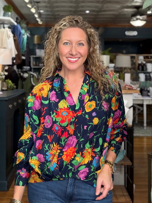 A smiling woman with curly blonde hair wearing a vibrant, multicolored floral print blouse with a dark navy base, paired with blue jeans, standing inside a retail store.