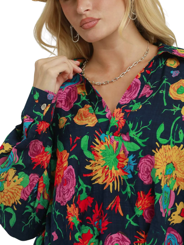 A close-up of a woman wearing a long-sleeve V-neck blouse with a vibrant multicolor floral print on a dark navy background. She is wearing a silver chain necklace and hoop earrings, and her blonde hair is visible against a plain white background.