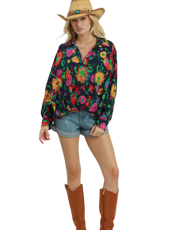A woman modeling a long-sleeve button-up blouse with a vibrant multicolor floral print on a dark background, paired with denim shorts, a straw cowboy hat, and brown boots against a plain white background.