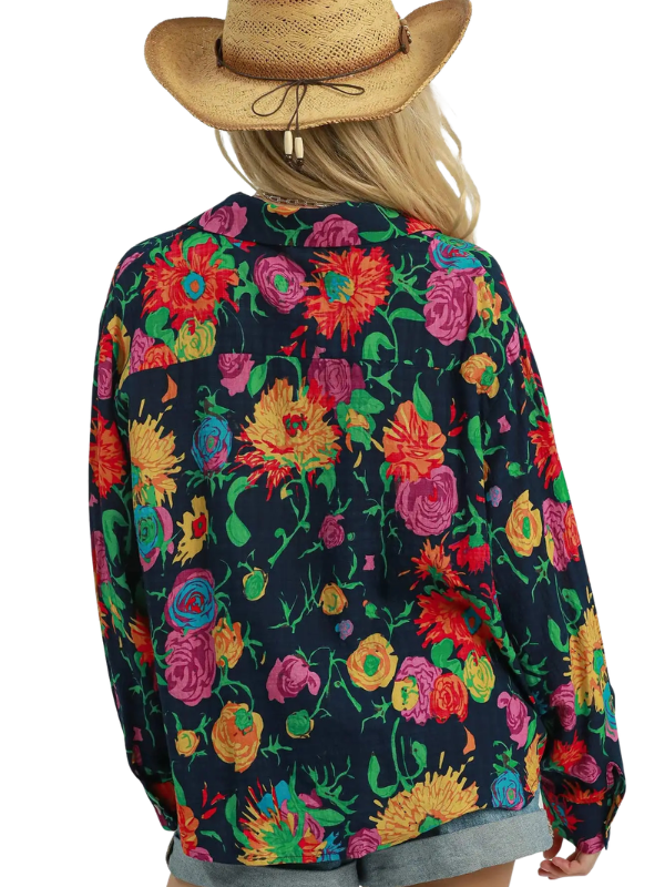 A woman shown from the back wearing a dark navy floral long-sleeve shirt with a vibrant multicolor print of flowers, paired with a straw cowboy hat and gray shorts, against a plain white background.