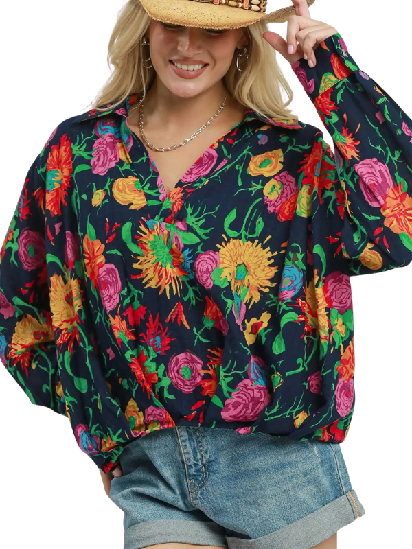 A woman wearing a long-sleeve V-neck blouse with a vibrant multicolor floral print on a dark navy background, paired with denim shorts and a straw hat against a plain white background.