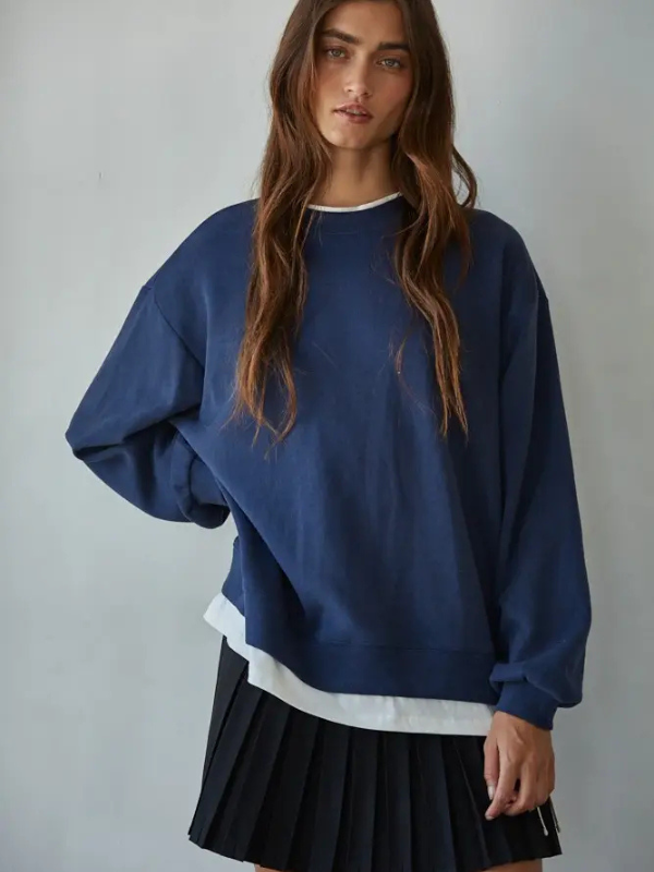 Woman wearing a blue sweatshirt over a white shirt with a black pleated skirt on a plain background