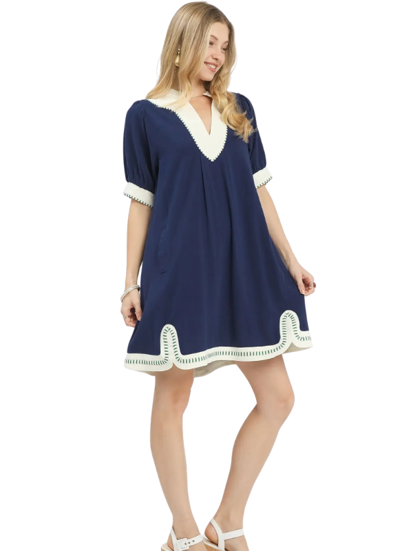 A woman modeling a short-sleeve navy blue dress with contrasting cream trim around the V-neck, sleeve cuffs, and hem, set against a plain white background.