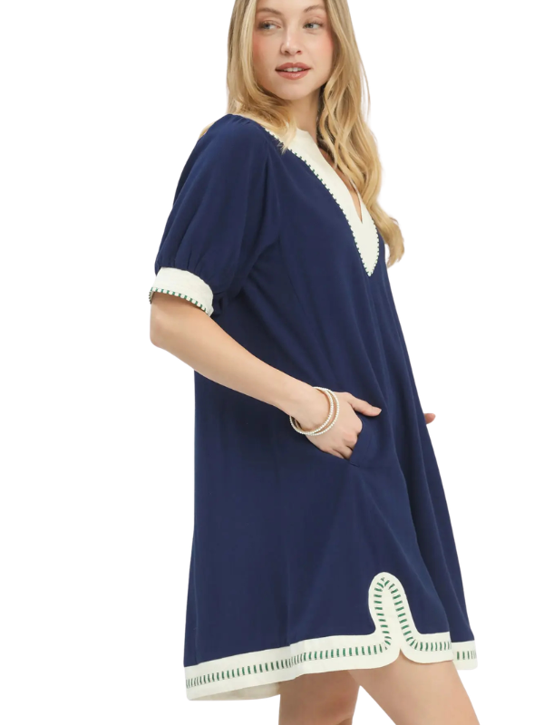 A woman modeling a navy blue tunic dress with cream-colored trim on the neckline, sleeve cuffs, and hem, set against a plain white background.
