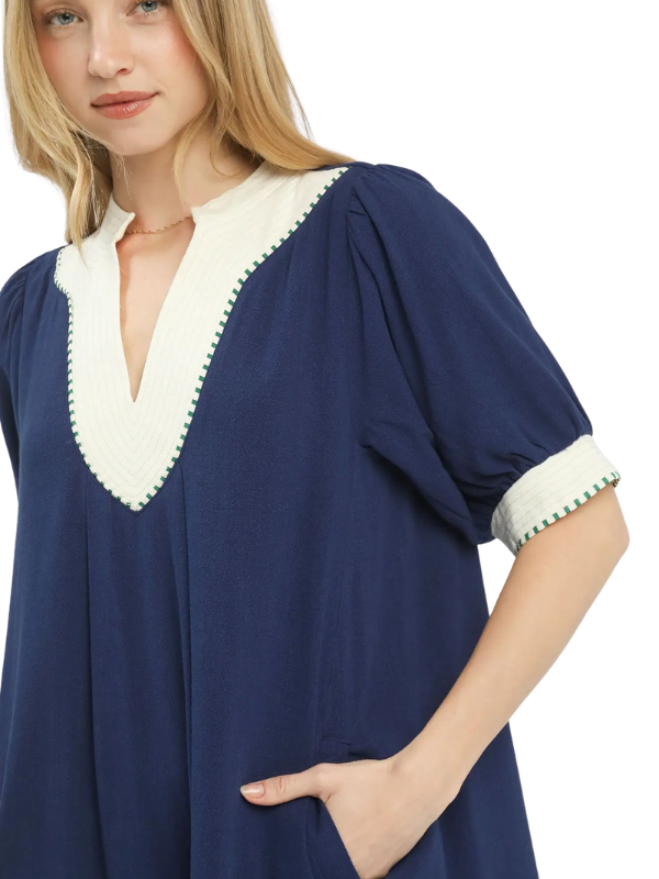 A woman wearing a loose navy blue tunic dress with a contrasting cream V-neck and sleeve cuffs, accented with green pom-pom trim, shown against a plain white background.