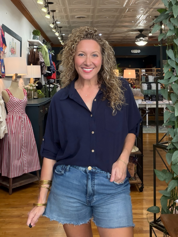 A smiling woman with curly blonde hair stands inside a boutique shop wearing a navy blue button-down shirt and light-wash denim shorts.