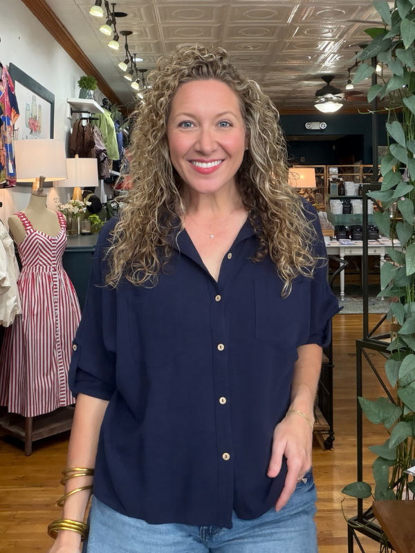 A smiling woman with curly blonde hair wearing a navy blue button-down shirt and light blue jeans, standing inside a clothing boutique.