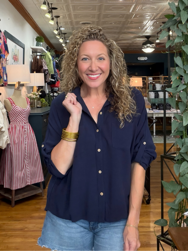 A smiling woman with curly blonde hair wearing a navy blue button-down shirt and light blue denim shorts, standing inside a retail boutique.