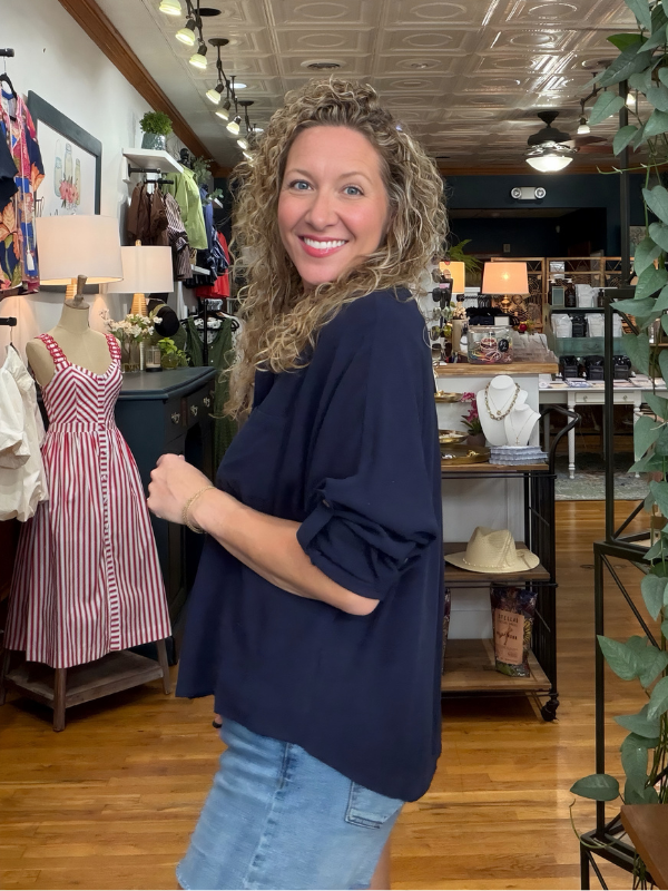 A woman with curly blonde hair smiling in a boutique, wearing a loose navy blue long-sleeve top and light wash blue jeans. The shop has wooden floors, a decorative ceiling, and clothing displays in the background.