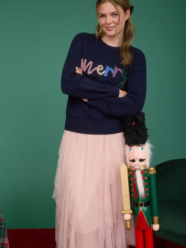 Woman wearing a navy sweater with colorful text, standing next to a nutcracker against a green background