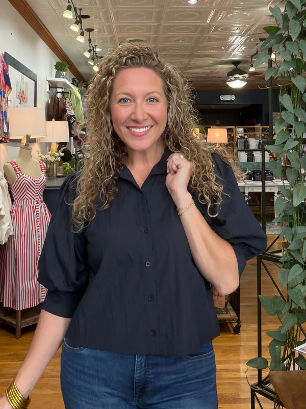 A smiling woman with curly blonde hair wearing a navy blue short-sleeve button-up shirt and blue jeans, standing inside a retail store with clothing racks, plants, and wood floors visible in the background.