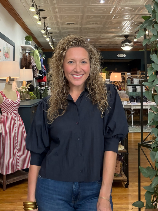 A smiling woman with curly blonde hair stands in a boutique shop wearing a navy blue puff-sleeve button-up shirt and blue jeans.